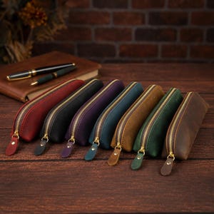 May include: A collection of seven rectangular pencil cases made of matte leather in red, black, purple, blue, tan, green, and brown. Each case features a gold-toned metal zipper closure with a matching leather pull tab, arranged in a row.