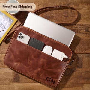 Op de afbeelding: Bruine leren laptoptas met schouderriem, met een laptop, telefoon, portemonnee en oplader. De tas heeft de initialen "C/M" op de voorkant. Op een houten oppervlak.