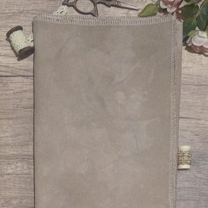 Puede incluir: Una pieza rectangular de tela beige con una textura tejida visible y bordes rematados. La tela está acompañada por unas tijeras de metal oscuro, un pequeño carrete de madera con encaje crema y un segundo carrete con adorno decorativo.