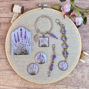 Puede incluir: Una colección de accesorios con temática de lavanda que incluye un llavero de metal con un dije cuadrado y una hoja, dos cabujones circulares, un dije de cuentas y una tarjeta de presentación impresa con dos orificios.