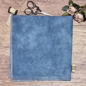 Puede incluir: Una pieza cuadrada de tela azul moteada con un patrón de cuadrícula visible y bordes rematados. La tela se acompaña de unas tijeras de metal ornamentadas, un pequeño carrete de encaje crema y un pequeño carrete de hilo de madera.