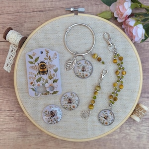 Puede incluir: Una colección de accesorios con temática de abejas que incluye una tarjeta rectangular, un llavero circular de metal con un dije de hoja, dos cabujones redondos y dos cordones con cuentas amarillas y verdes, todos con ilustraciones florales.
