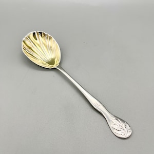 Pode incluir: Uma colher de prata com uma tigela dourada e um cabo longo e fino. A tigela tem um design canelado e o cabo apresenta um padrão de folhas gravadas. A colher é provavelmente para servir ou comer.