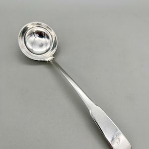 May include: A silver ladle with a round bowl and a long, slender handle. The handle has a slightly flattened shape with a small, decorative marking near the end. The ladle is made of a shiny, reflective metal.