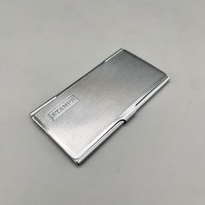 May include: A rectangular, silver-colored stamp case with a textured surface. The word "STAMPS" is embossed on the front. The case has a hinged design and a polished finish.