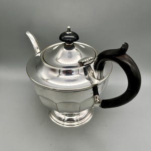Op de afbeelding: Een zilveren theepot met een gepolijste metalen afwerking, voorzien van een gefacetteerd lichaam en een voetstuk. De pot heeft een scharnierend deksel met een kleine zwarte knop, een gebogen zwart handvat en een slanke zilveren tuit.