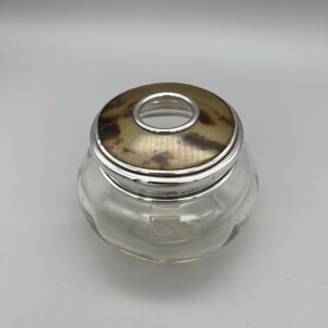 May include: A clear glass jar with a silver rim and a decorative lid. The lid has a gold-colored center with a circular opening. The jar has a rounded shape and sits on a plain background. This jar is suitable for storing small items.