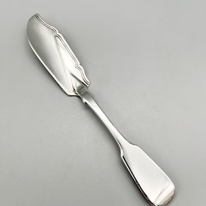 Puede incluir: Un cuchillo de mantequilla de plata con un borde decorativo festoneado y un mango redondeado.