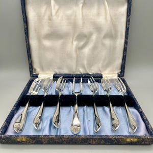 May include: A set of silver forks in a blue and gold patterned box. The forks are arranged neatly on a blue satin lining, with a black velvet divider. Each fork has a decorative floral design on the handle.