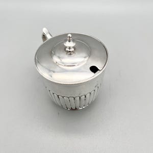 May include: A polished silver mustard pot with a lid featuring a small finial knob. The cylindrical body has vertical fluted detailing along the lower section and a beaded rim. A small notch is cut into the lid to accommodate a spoon.