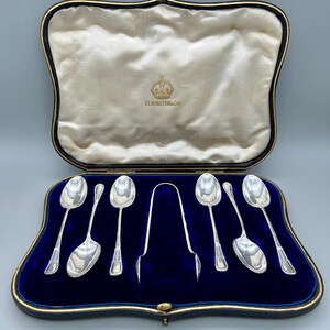 Könnte beinhalten: Ein Set aus silbernen Löffeln und Zangen in einem blau samtgefütterten Etui mit goldenem Rand. Der Innenraum ist aus cremefarbenem Satin. Das Etui trägt die Aufschrift "ELKINGTON & Co.", und die Löffel sind ordentlich angeordnet.