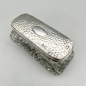 May include: A rectangular, silver-toned box with a textured, hammered surface on the lid and a smooth, oval accent. The base is clear, faceted glass. The box is likely for jewelry or small items.