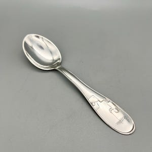 May include: A silver spoon with a rounded bowl and a long, slender handle. The handle features an engraved design near the end. The spoon is made of a shiny, reflective metal.