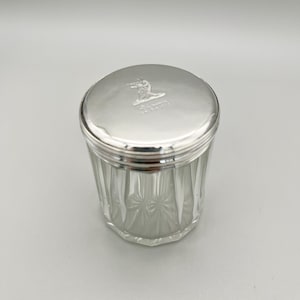 May include: A clear glass jar with a silver lid. The jar has a ribbed design on the sides and a starburst pattern on the bottom. The lid is silver with an embossed design. The jar is likely for storing small items.