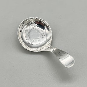 May include: A small, silver spoon with a round bowl and a short handle. The bowl has a decorative, engraved design in the center. The handle also features an engraved pattern. The spoon is likely made of metal and has a shiny finish.