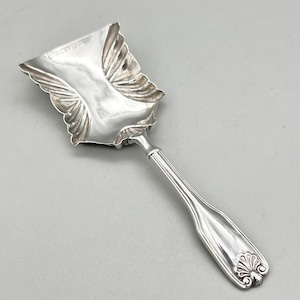 May include: A silver serving scoop featuring a wide, rectangular bowl with scalloped, shell-like edges and a polished finish. The handle is slender and straight, terminating in a decorative shell motif at the base, with hallmarks visible on the bowl.
