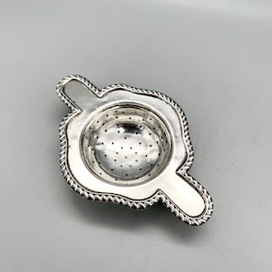 May include: A silver tea infuser with a decorative rope-like border. The infuser has a round, perforated bowl for tea leaves and two small handles. The tea strainer is designed to sit atop a teacup.