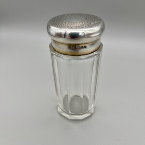 May include: A clear, faceted glass jar with a silver-colored lid. The lid has a detailed, circular pattern. The jar appears to be a vintage item, possibly for storing perfume or other small items. The jar is on a plain background.