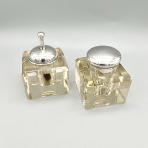 May include: Two clear glass inkwells with silver lids. The inkwells are square shaped and have a small opening in the center. The lids are round and have a small knob on top.