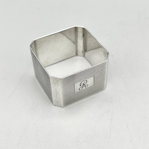 May include: A silver napkin ring with a geometric design. The ring is square with a textured pattern on the sides and a rectangular panel with the letter "R" engraved on it. The napkin ring is made of a shiny, reflective metal.