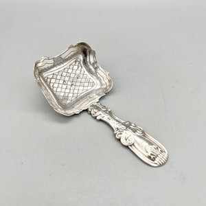 Puede incluir: Una cuchara de azúcar de plata con un cuenco cuadrado y un mango adornado. El cuenco tiene un patrón decorativo y el mango tiene un diseño de desplazamiento.