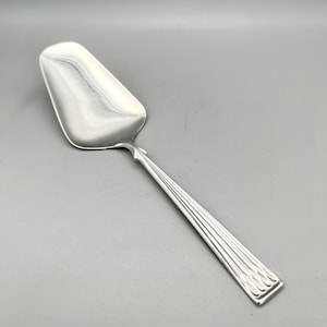 May include: A silver pie server with a wide, rounded blade and a long, rectangular handle. The handle features a series of parallel lines and a decorative pattern at the end. The server is made of a shiny, reflective metal.
