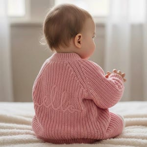 May include: A soft, chunky knit jumper in a dusty pink colour featuring long sleeves and a ribbed texture. The back of the garment displays the word hello embroidered in a matching pink yarn, with a classic crew neckline and a fitted hem.