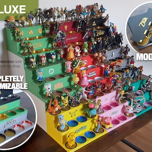 Puede incluir: Un expositor modular de varios niveles para figuras de videojuegos. El expositor está hecho de madera y tiene una variedad de colores, incluyendo verde, rojo, amarillo y rosa. El expositor está diseñado para sostener una variedad de figuras, incluyendo personajes de las franquicias Zelda, Mario y Pokémon. El expositor también es personalizable, con la posibilidad de agregar niveles y estantes adicionales. El texto "DELUXE", "COMPLETELY CUSTOMIZABLE", "ZELDA", "LINK", "SUPER MARIO", "MODULAR" es visible en el expositor.