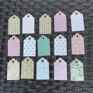 May include: A collection of fifteen decorative gift tags with scalloped edges. The tags feature various patterns, including stripes, floral designs, and geometric shapes, in pastel colors like pink, green, and blue. Each tag has a reinforced eyelet for attaching a string.