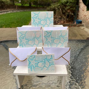 May include: A set of six white note cards with a blue and white seashell design. The cards are stacked on a white wooden stand. The stand is on a glass table.