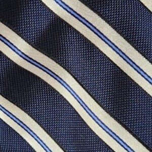 May include: A necktie featuring a diagonal striped pattern. The design consists of wide, textured dark navy blue bands alternating with narrower cream-colored stripes, each centered with a thin, vibrant light blue line running through the middle.