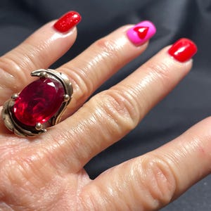 Può includere: Un anello argentato con una grande gemma rosso scuro a taglio ovale. L'anello presenta un motivo decorativo a foglie sui lati della gemma. Le unghie sono dipinte in tonalità rosse e rosa.