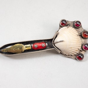 May include: A unique silver brooch featuring a seashell, red accents, and seven red gemstones. The brooch has a pen nib with "C-4" engraved on it. The design is intricate and artistic.