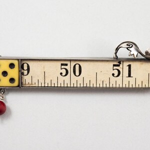 May include: A unique, handcrafted pendant featuring a vintage measuring tape with the numbers 9, 50, and 51. A yellow die with six black dots is attached to the left, and a red bead hangs below. A silver floral design adorns the top.