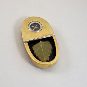 K&ouml;nnte beinhalten: Ein ovaler Anh&auml;nger aus Gold mit einem kleinen runden Fenster, das ein schwarzes Kreuz zeigt. Der Anh&auml;nger l&auml;sst sich &ouml;ffnen und zeigt ein gepresstes gr&uuml;nes Blatt.