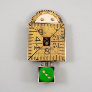 May include: A gold and silver-toned metal brooch with a ruler design and a green die with gold dots. The ruler has numbers 4, 8, 12, 2, 24, and 44.