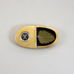 May include: A gold and black oval-shaped brooch with a leaf design and a black and white 'X' in a circle.