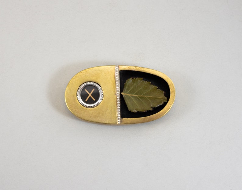 K&ouml;nnte beinhalten: Eine ovale Brosche in Gold und Schwarz mit einem Blattdesign und einem schwarzen und wei&szlig;en 'X' in einem Kreis.