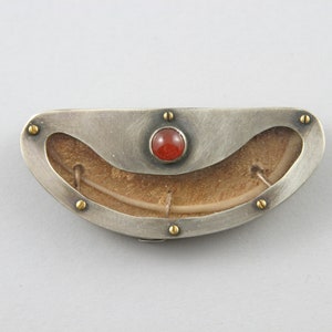Könnte beinhalten: Eine Brosche aus Silber und Gold mit einer geschnitzten, hellbraunen, organischen Form im Inneren. Die Brosche hat einen roten Karneol-Stein in der Mitte.