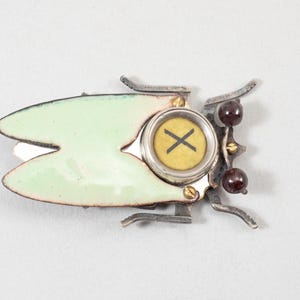 May include: A unique, handcrafted brooch in the shape of a stylized insect. The brooch features a pale green enamel body, silver-toned metal accents, and a central circular element with a yellow background and a black "X" symbol. Two dark red beads add a pop of color.