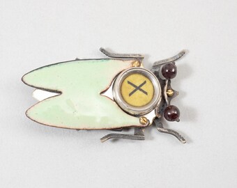 Könnte beinhalten: Einzigartige, handgefertigte Brosche in Form eines stilisierten Insekts. Die Brosche hat einen hellgrünen Emaille-Körper, silberfarbene Metallakzente und ein zentrales kreisförmiges Element mit gelbem Hintergrund und einem schwarzen "X"-Symbol. Zwei dunkelrote Perlen setzen einen Farbakzent.