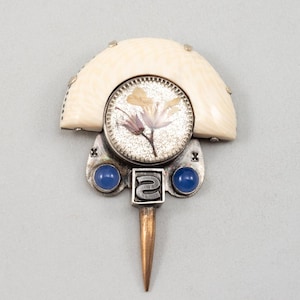 May include: A unique brooch featuring a cream-colored, semi-circular top. A circular, silver-toned element showcases a pressed flower design. Two blue gemstones flank a silver-toned square with a stylized design, and a copper-colored pin extends downward.