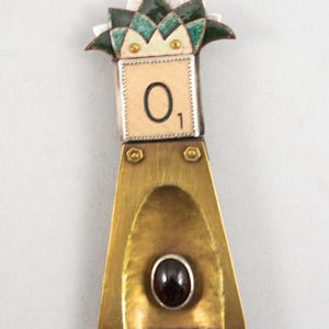 May include: A brass and silver brooch with a green and white enamel flower design, a tile with the number "01" and a dark red gemstone.