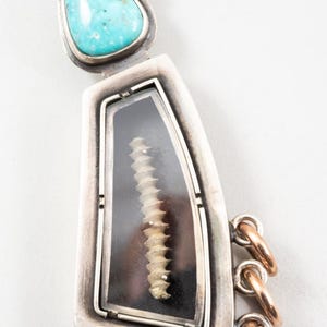 May include: A silver pendant with a turquoise stone and a clear window showcasing a beige, spiral-shaped object. The pendant has gold accents and three gold rings attached.