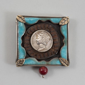 May include: A silver coin with a woman's profile on it is set in a square frame with a turquoise blue background. The frame is made of metal and has a brown, textured border. There are gold leaf accents on the corners of the frame. A red bead hangs from the bottom of the frame.