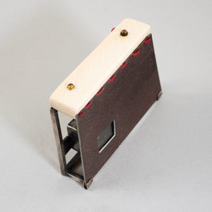 May include: A small, rectangular box made of metal and wood. The box has a brown leather exterior with red stitching and a white wood top. The top has two brass screws. The box has a small window on the front.