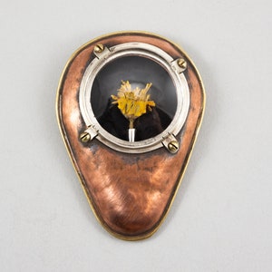 May include: A small, teardrop-shaped copper and brass pendant with a round glass window. Inside the window is a dried yellow flower with a black background.