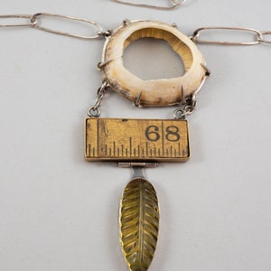 Puede incluir: Un collar de cadena de plata con un gran colgante redondo de color blanquecino con un agujero en el centro. Colgando del colgante hay una pequeña regla de madera rectangular con el número "68" impreso en ella. Debajo de la regla hay un pequeño colgante en forma de hoja amarilla.