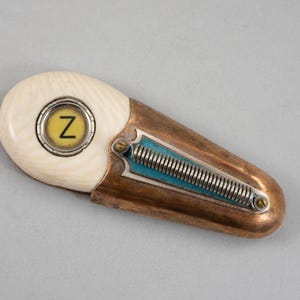 May include: A small, oval-shaped, ivory-colored object with a copper-colored casing. The ivory portion has a circular cutout with a yellow background and the letter "Z" in black. The copper casing has a blue and silver spring-like design.