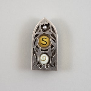 May include: A silver pendant with an ornate design, featuring a yellow circle with the letter "S" inside, a white shell, and a small pearl.